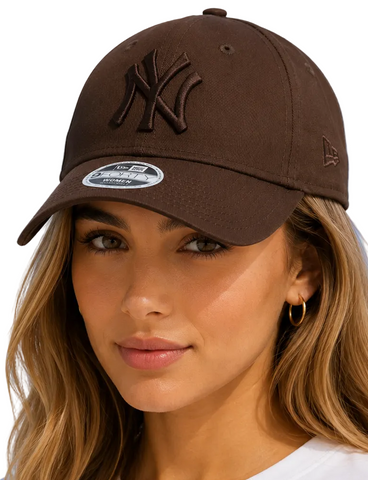 A woman wearing a dark brown New Era New York Yankees baseball cap, photographed against a bright blue sky with light clouds.