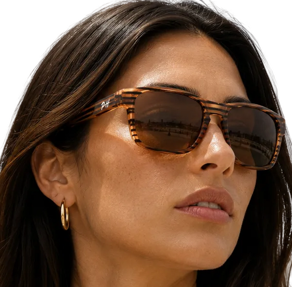 A close-up of a woman wearing rectangular tortoiseshell sunglasses with dark brown lenses and gold hoop earrings, shown against a plain white background.